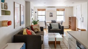 Uma sala de estar moderna com sofá, mesa de centro e área de jantar com mesa e cadeiras. Há quadros nas paredes, uma planta e um armário de madeira.