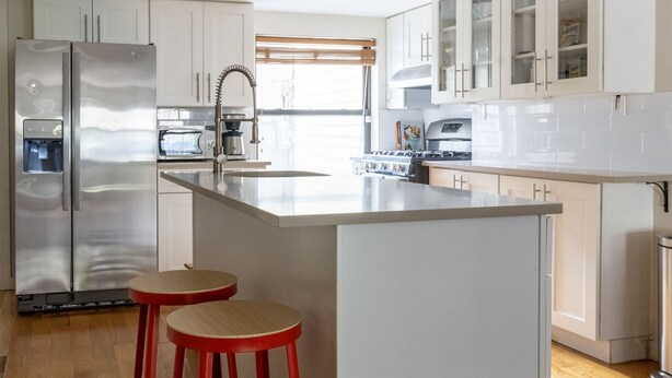 Uma cozinha moderna com armários brancos, uma geladeira de aço inoxidável, uma ilha central com banquetas e piso de madeira.