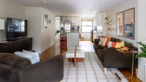 Uma sala de estar moderna com um sofá, uma mesa de centro e uma televisão. Dá para ver uma cozinha ao fundo, com uma geladeira e uma janela.