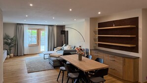 Un espacio habitable moderno con comedor, sofá, cocina con estantes empotrados y un amplio ventanal con cortinas.