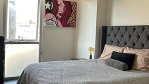 Une chambre avec un grand lit, une tête de lit capitonnée et une vue sur le paysage urbain par la fenêtre.