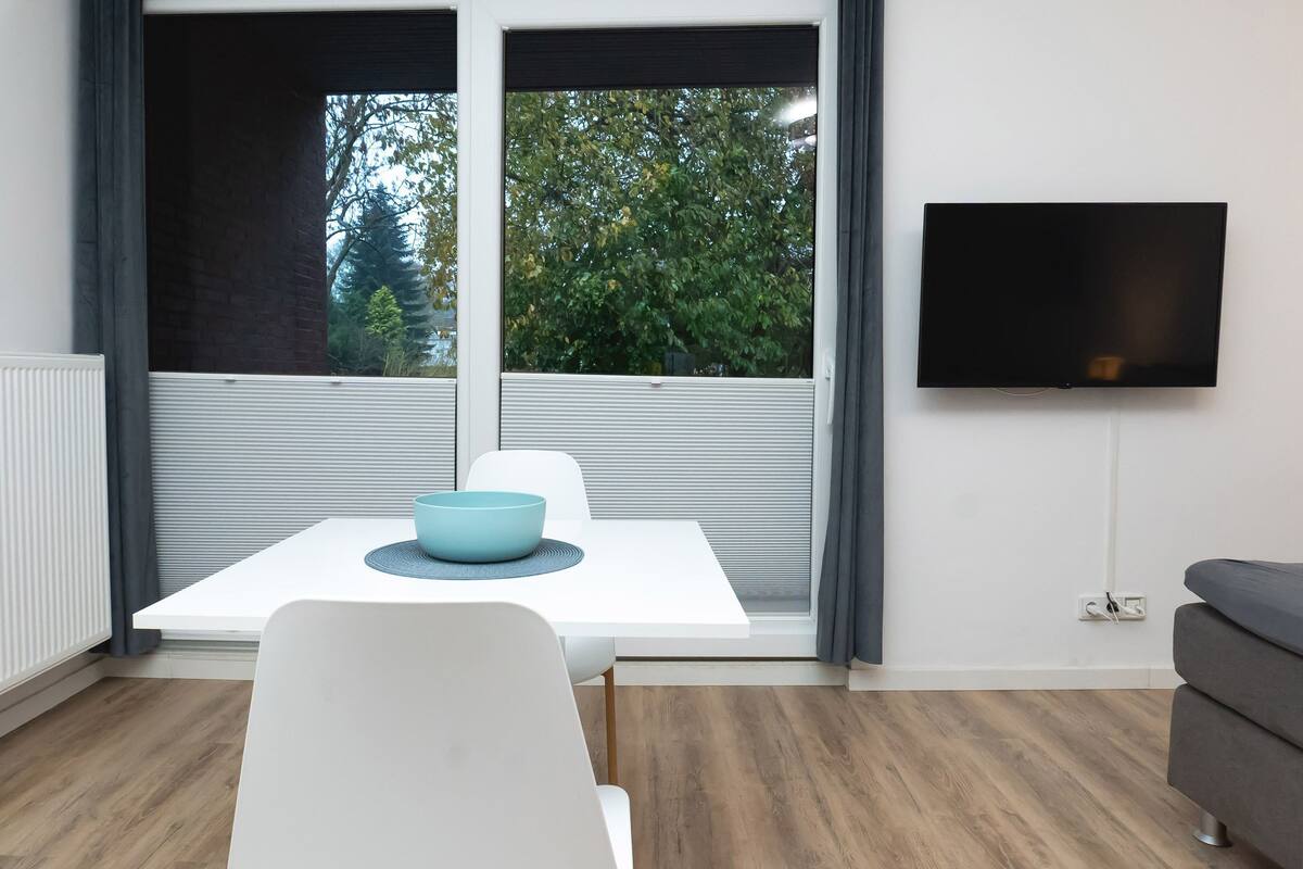 Une pièce moderne avec une table blanche, un bol bleu, un canapé gris et un téléviseur fixé au mur.
