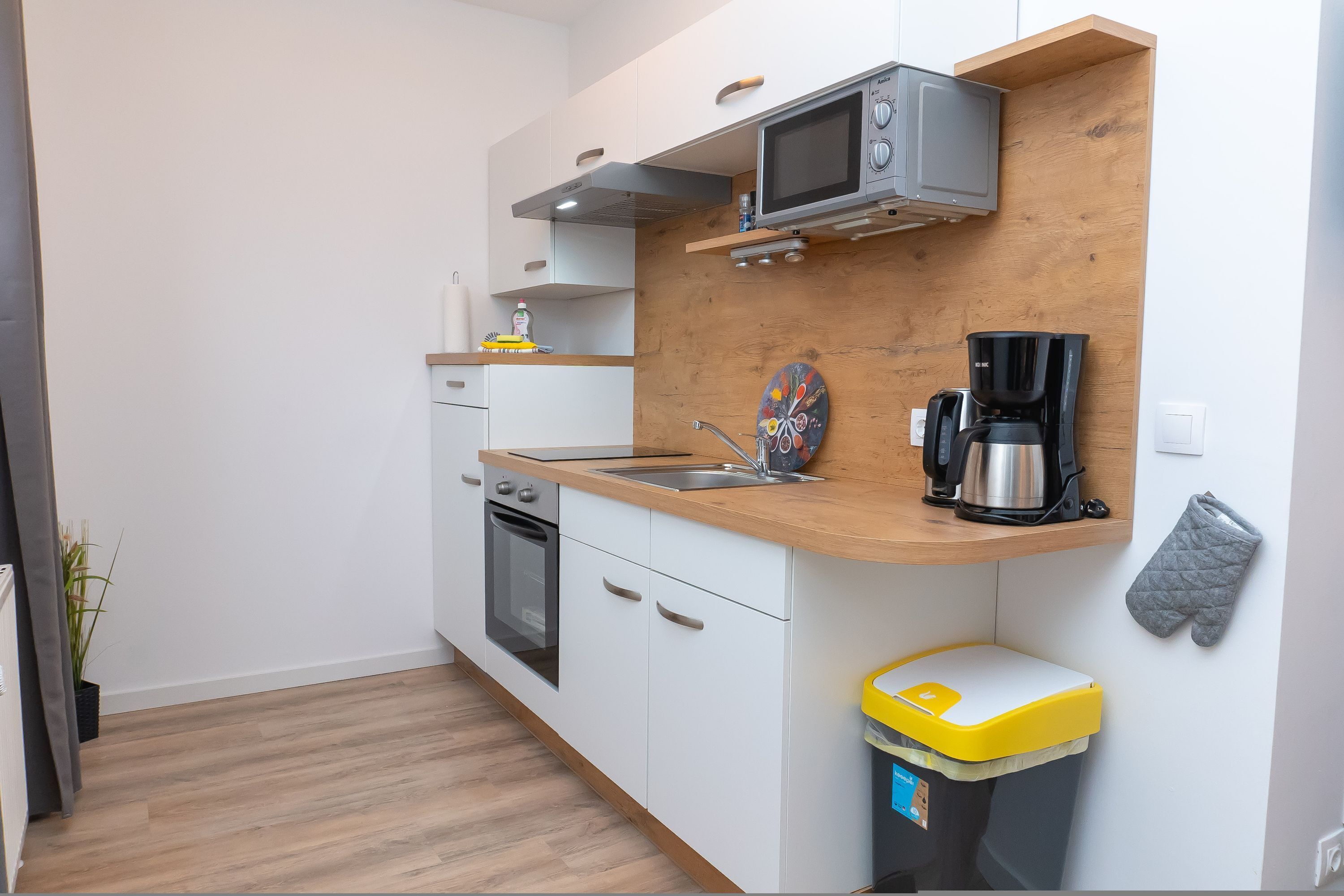 Una cocina compacta con armarios blancos, encimera de madera, microondas, cafetera y un cubo de basura.