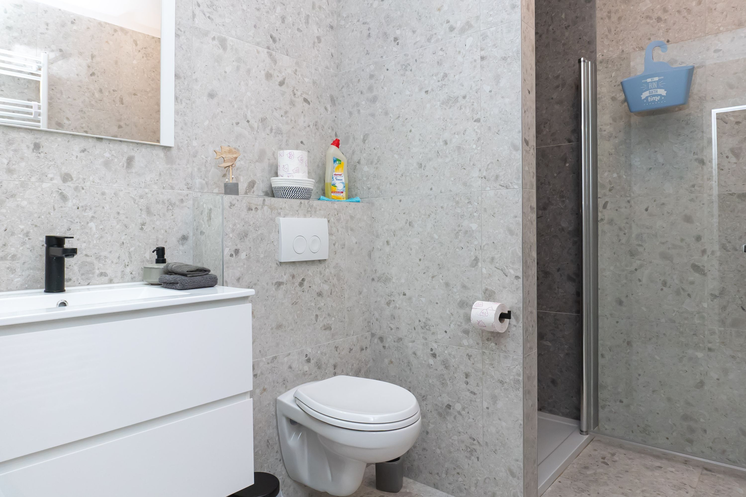 Un baño moderno con lavamanos, inodoro y ducha blancos. Hay artículos de aseo en la pared y un bolso azul colgado en la puerta de la ducha.