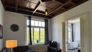 A living room with a sofa, a coffee table, a TV, a lamp, a clock, and a window with curtains. - Infinity Stay: Nordsee Apartments (Wilhelmshaven)