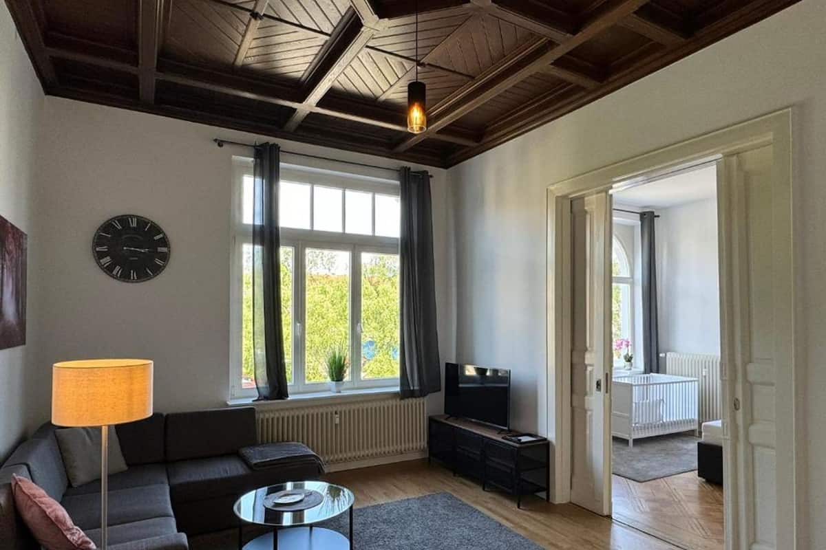 Un salon comprenant un canapé, une table basse, un téléviseur, une lampe, une horloge et une fenêtre avec des rideaux.