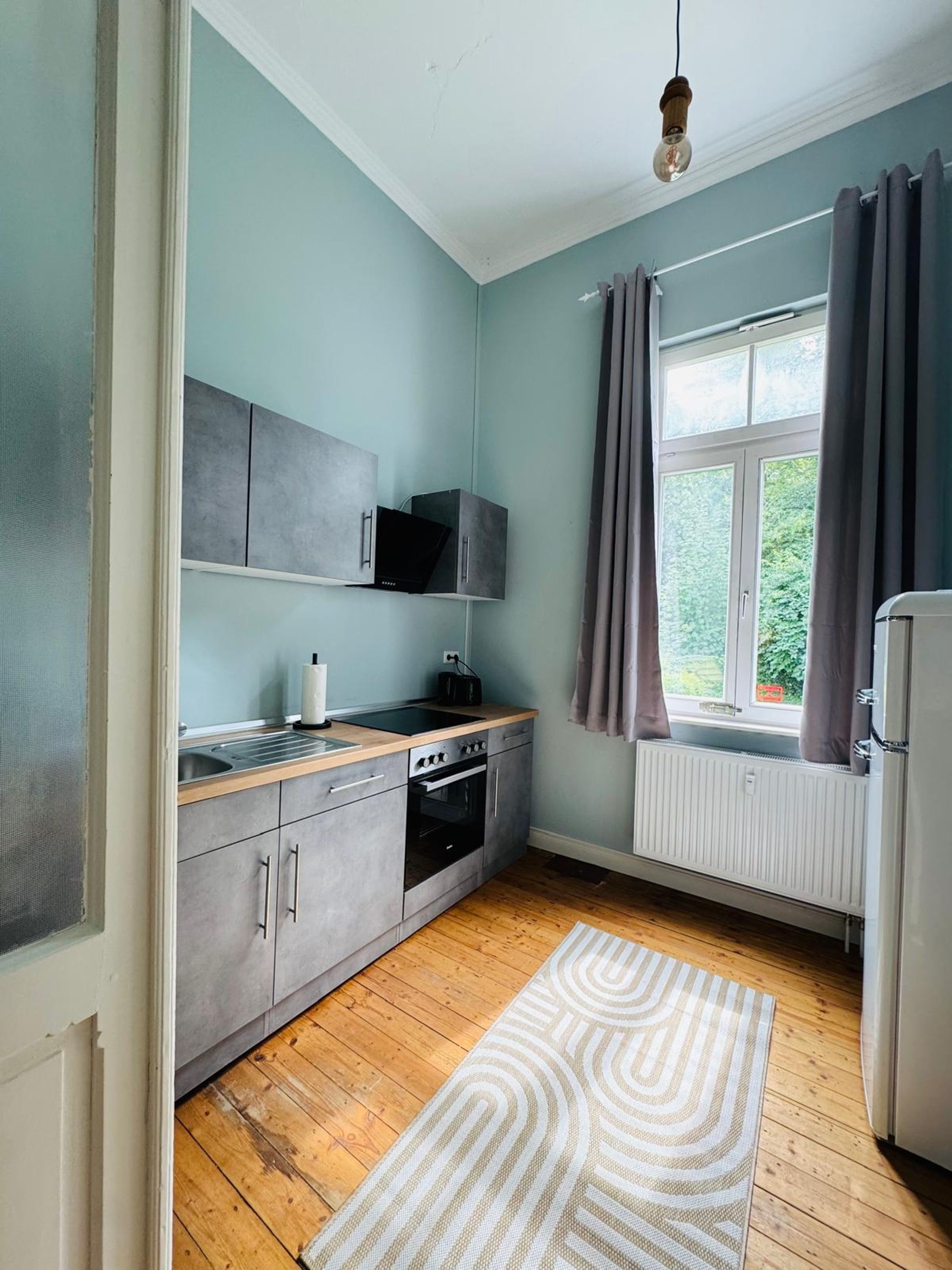 Une cuisine compacte avec des meubles gris, un plan de travail en bois, un four encastré et une fenêtre avec des rideaux.