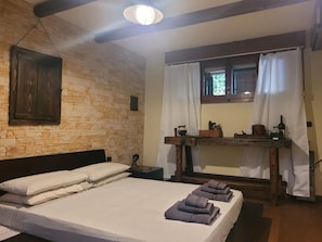 Ein Zimmer mit einem Bett, einem Holz-Kopfstück, einer Steinwand, einem Holztisch mit Spiegel und einem Fenster mit Vorhängen.
