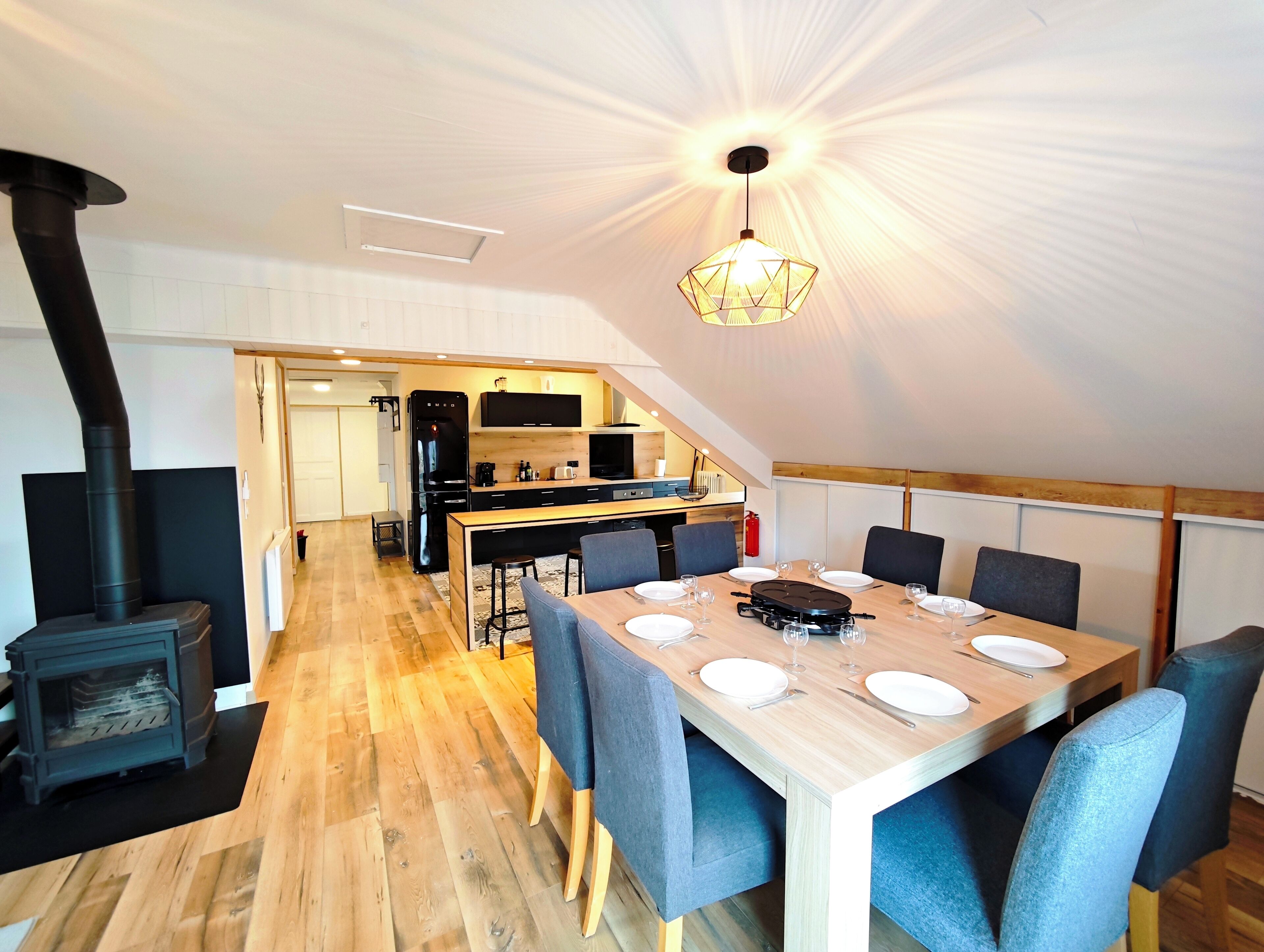 Une cuisine moderne dotée d’une cuisinière intégrée, d’un coin repas avec une table en bois et des chaises bleues, et d’un plafond haut agrémenté d’un luminaire original.