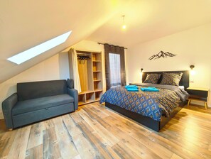 Een slaapkamer met een schuine kap, een inbouwkast, een bed met behang en een grijze bank.