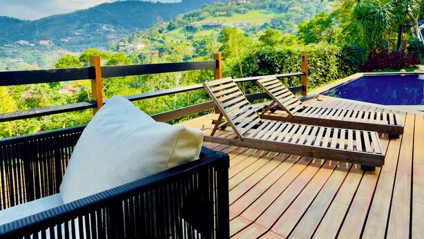 A wooden deck with lounge chairs and a pool, overlooking a lush landscape with hills and trees.
