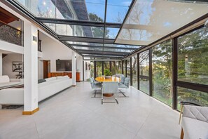 A spacious living area with a glass ceiling, a white sofa, a dining table with chairs, and a TV mounted on the wall.