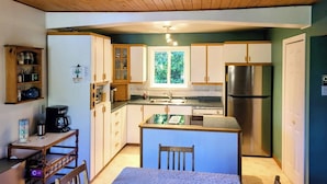A compact kitchen with wooden cabinets, a stainless steel refrigerator, and a window providing natural light. - Cozy Nature Cottage Retreat – Relax, Unwind & Recharge (Notre-Dame-de-Pontmain)