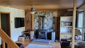 A cozy living room with a stone fireplace, wooden ceiling, and a bookshelf. - Cozy Nature Cottage Retreat – Relax, Unwind & Recharge (Notre-Dame-de-Pontmain)