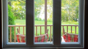 A window with three panes, each offering a view of a wooden deck with red plaid chairs and a lush green landscape. - Cozy Nature Cottage Retreat – Relax, Unwind & Recharge (Notre-Dame-de-Pontmain)