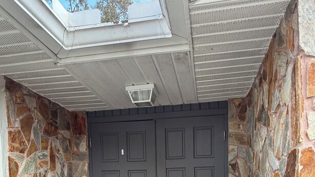 A dark grey double door with a keyhole and a lock, flanked by two large black planters with green shrubs, set against a stone wall.