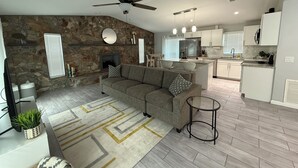 A modern living room with a stone wall, a fireplace, a grey sectional sofa, and a kitchen in the background.