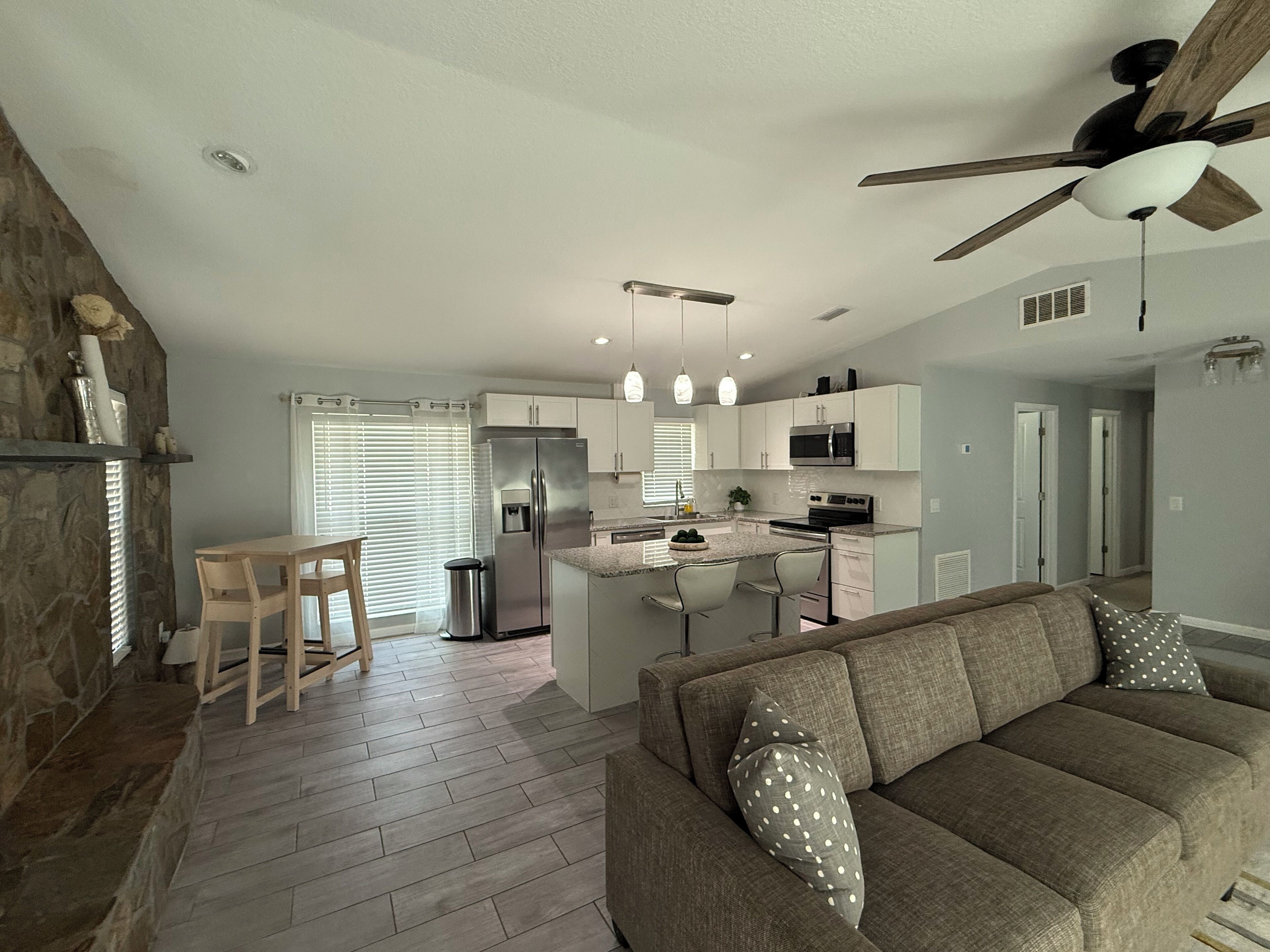 A modern kitchen with white cabinets, a stainless steel refrigerator, and a microwave. There is a dining area with a wooden table and chairs. A ceiling fan is visible above the kitchen.