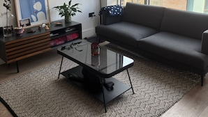 Ein modernes Wohnzimmer mit einem grauen Sofa, einem Glastisch, einer schwarzen Lampe und Blick auf eine Stadtlandschaft durch große Fenster.