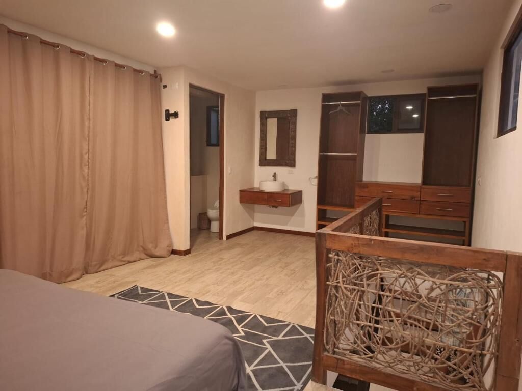 A bedroom with a bed, a wooden headboard, a bedside table with a mirror, a wardrobe, and a bathroom visible through an open door.
