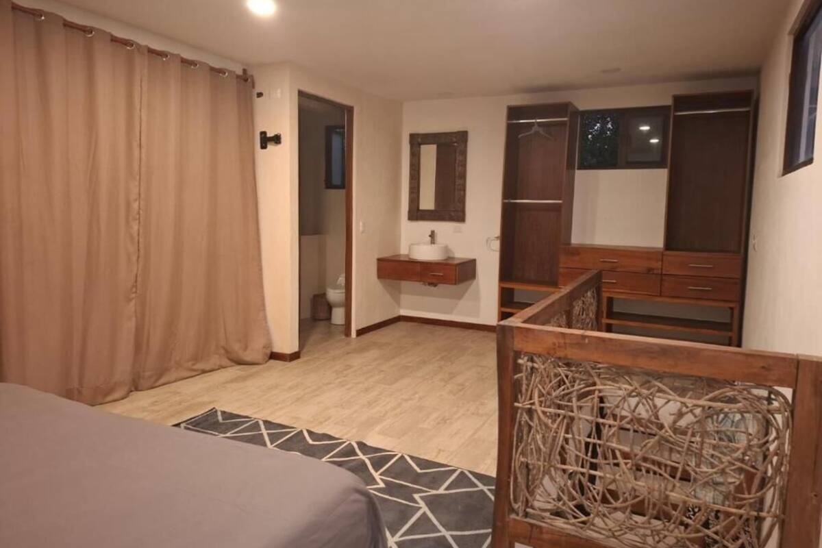 A bedroom with a bed, a wooden headboard, a bedside table with a mirror, a wardrobe, and a bathroom visible through an open door.