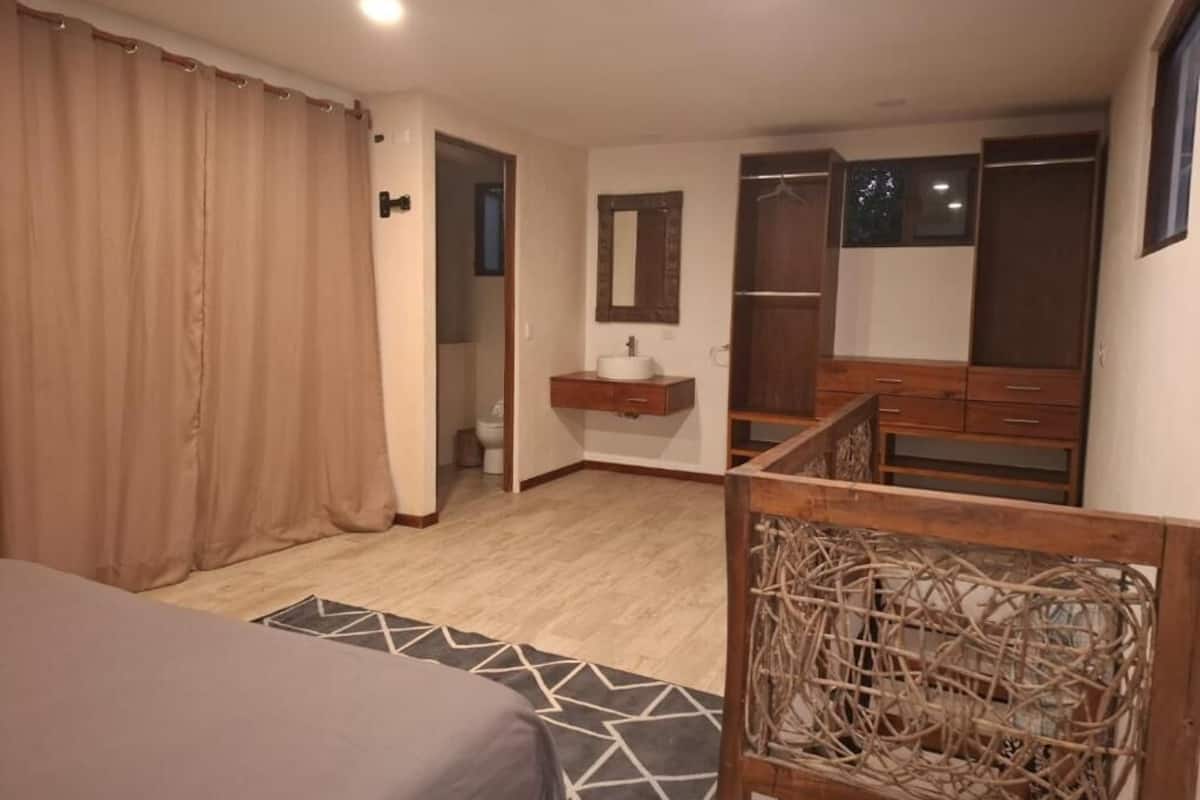 A bedroom with a bed, a wooden headboard, a bedside table with a mirror, a wardrobe, and a bathroom visible through an open door.