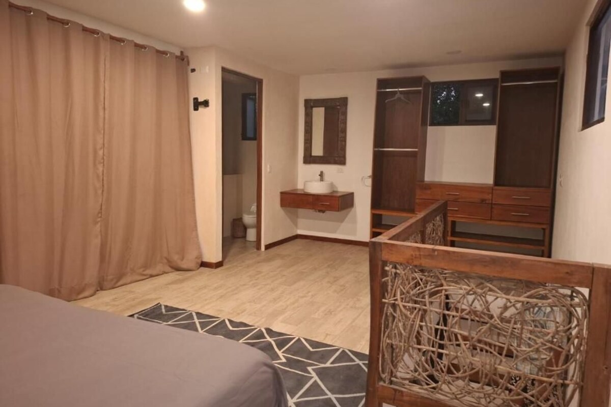Une chambre à coucher avec un lit, une tête de lit en bois, une table de chevet avec un miroir, une armoire et une salle de bain visible à travers une porte ouverte.