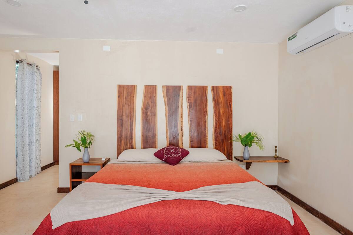 A bedroom with a wooden headboard, a red bedspread, two bedside tables with plants, and a white air conditioning unit on the wall.