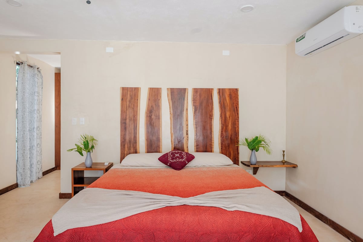 A bedroom with a wooden headboard, a red bedspread, two bedside tables with plants, and a white air conditioning unit on the wall.