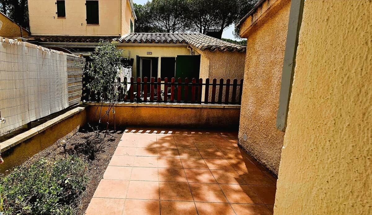 Un patio estrecho con suelo de baldosas, un pequeño jardín y un edificio con tejado de tejas y valla de madera.