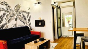 Un espacio habitable compacto con sofá, una pequeña mesa de madera, televisor y una zona de cocina con taburetes.