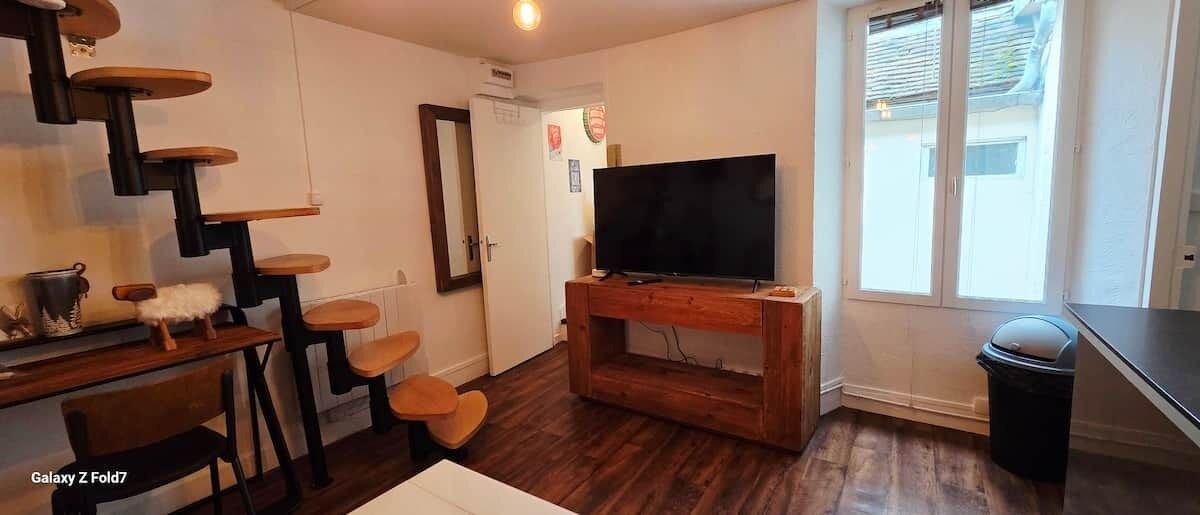 A compact living space with a staircase, a flat-screen TV on a wooden stand, a trash bin, and a window with blinds.