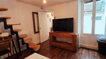 A compact living space with a staircase, a flat-screen TV on a wooden stand, a trash bin, and a window with blinds.