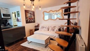 Un espace de vie compact comprenant un canapé, une table basse blanche et une chaise en bois. On aperçoit à l’arrière-plan un coin cuisine avec un four à micro-ondes et un évier.