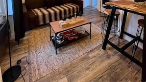 Een woonkamer met een bruine bank, een houten salontafel en een eethoek met een houten tafel en barstoelen.