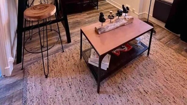 Een ruimtelijk ingerichte leefruimte met kitchenette, een eethoek met tafel en stoelen, een bank en een bijzettafeltje. Er is een raam met zonwering, een vloerlamp en een wandplank met decoratieve objecten.