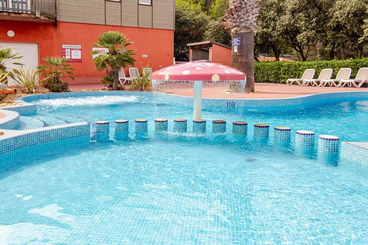 Une piscine dont le bord est revêtu de carrelage, un parasol rouge et des chaises longues.