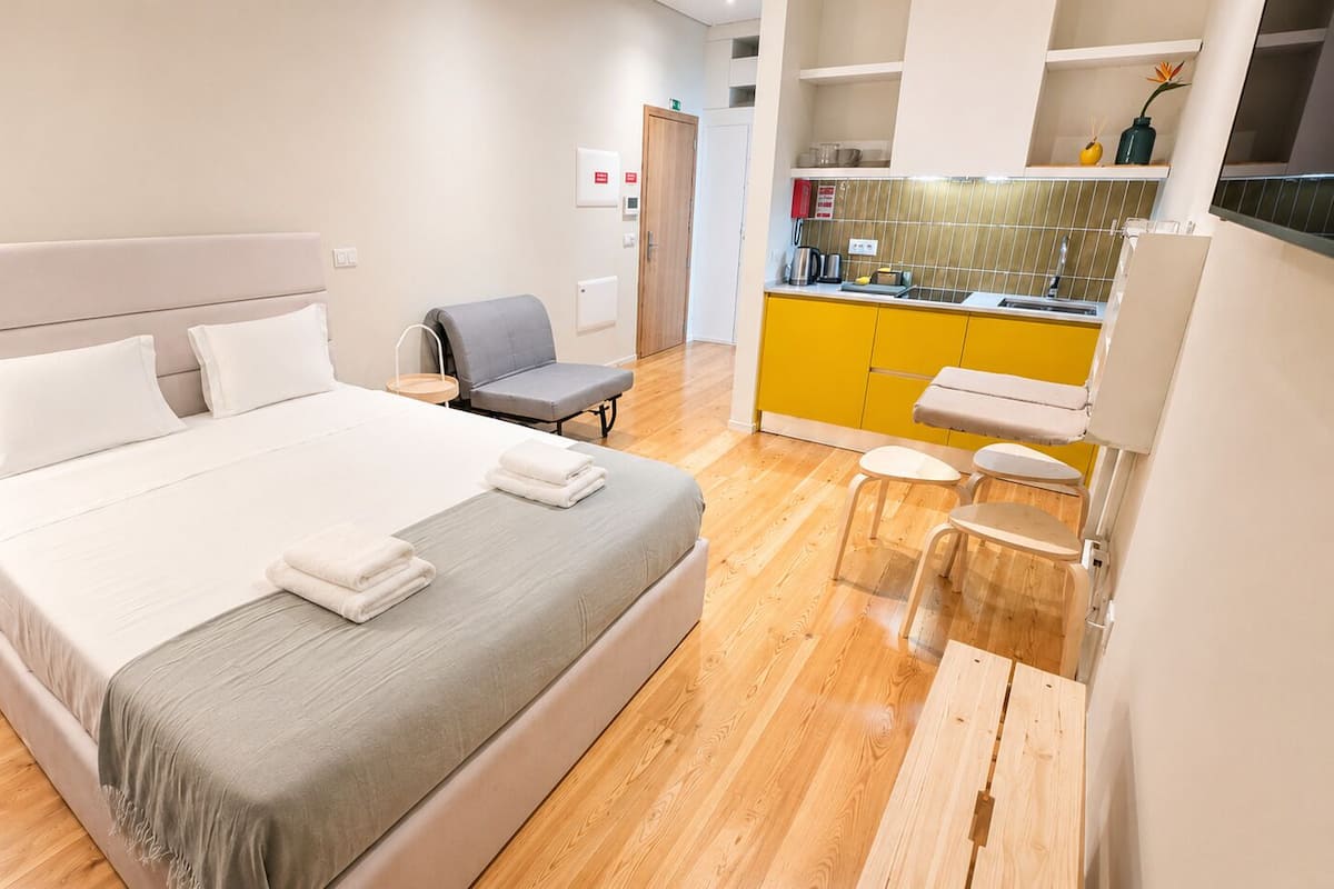 A hotel room with a bed, a chair, a small table, a kitchenette, and a bathroom.
