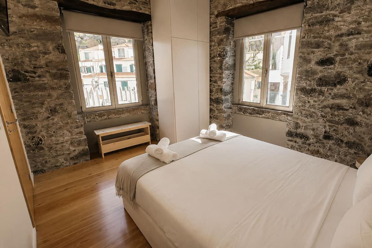 A bedroom with a stone wall, a wooden bench, a bed with white linens, and two windows with views of buildings.