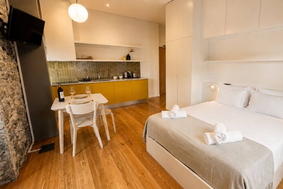 A hotel room with a bed, a dining table with chairs, a kitchen area, and a television mounted on the wall.