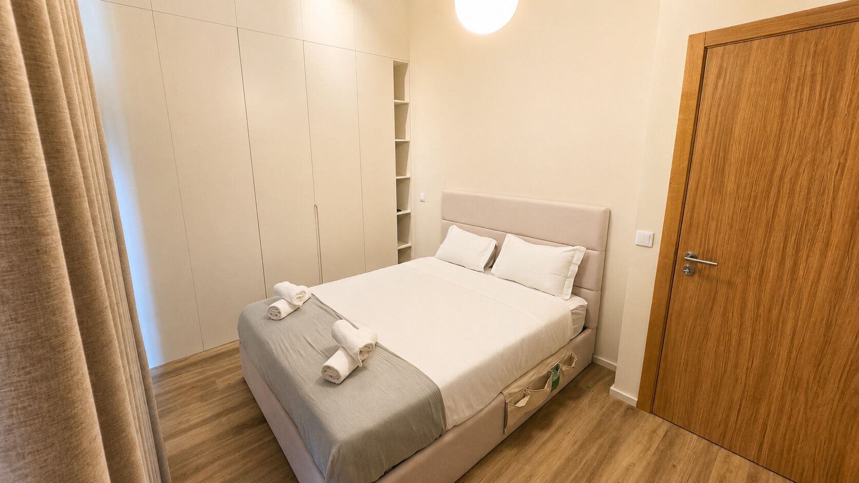 A neatly made bed with pillows and rolled towels, a wooden door, and a wardrobe with shelves.