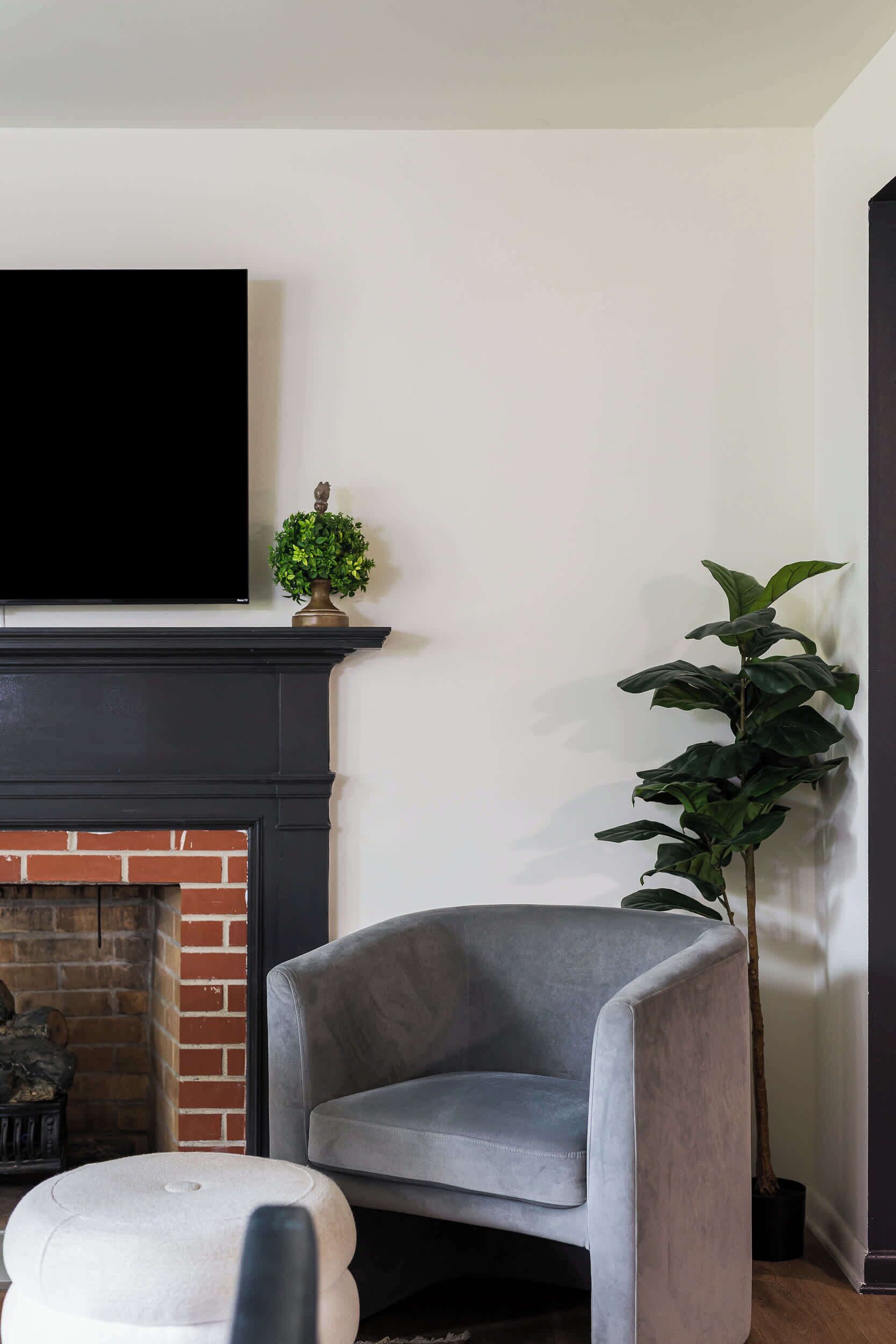 Un salon avec une cheminée, un fauteuil gris, un pouf blanc, une grande plante en pot et un téléviseur à écran plat fixé au-dessus de la cheminée.