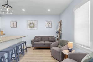A modern living room with a grey sofa, a small round table with a lamp, a bar area with stools, and a large window with blinds. - New Downtown 2bd w. Modern Southern Charm (Beaufort)