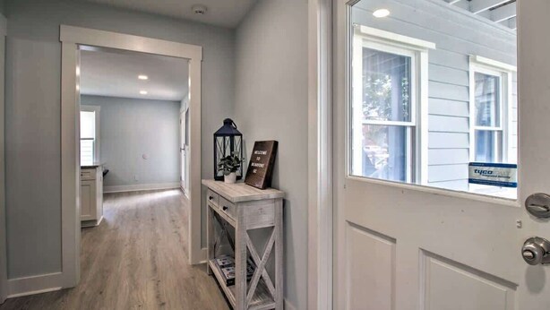 Un pasillo con piso de madera, una puerta blanca con ventana y una pequeña mesa con un cartel de bienvenida.
