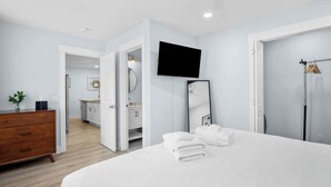 Una habitación de hotel moderna con una cama grande, un televisor de pantalla plana, un espejo, un aparador de madera y un baño visible a través de una puerta abierta.