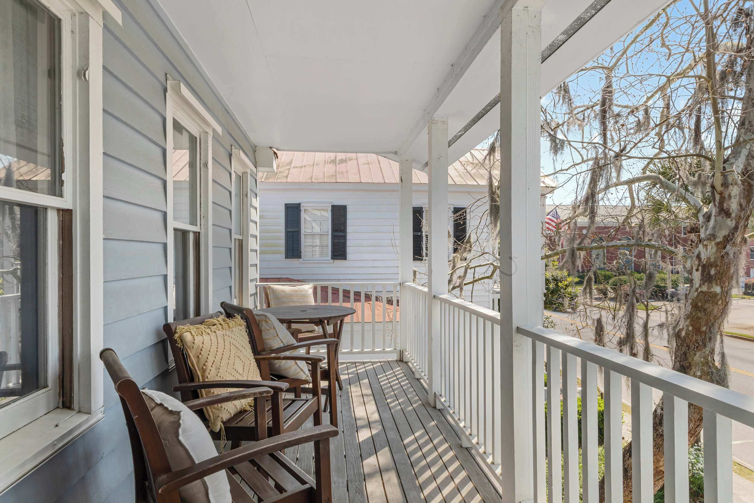 A porch with wooden chairs and a table, a white railing, and a view of a house and trees.