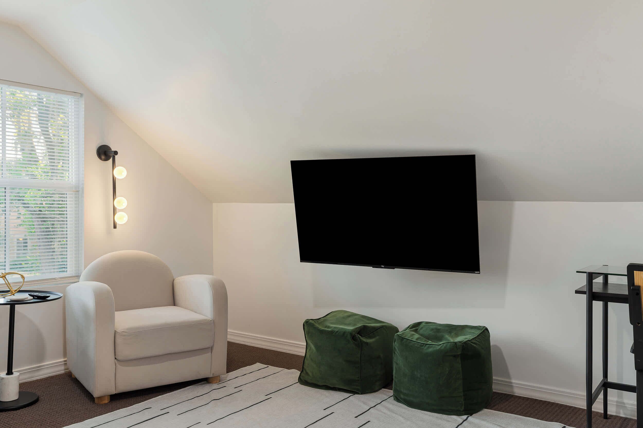 Ein Zimmer mit weißem Sessel, einem kleinen runden Tisch, einem grünen Pouf, einem flachbildfernseher an der Wand und einem Fenster mit Jalousien.