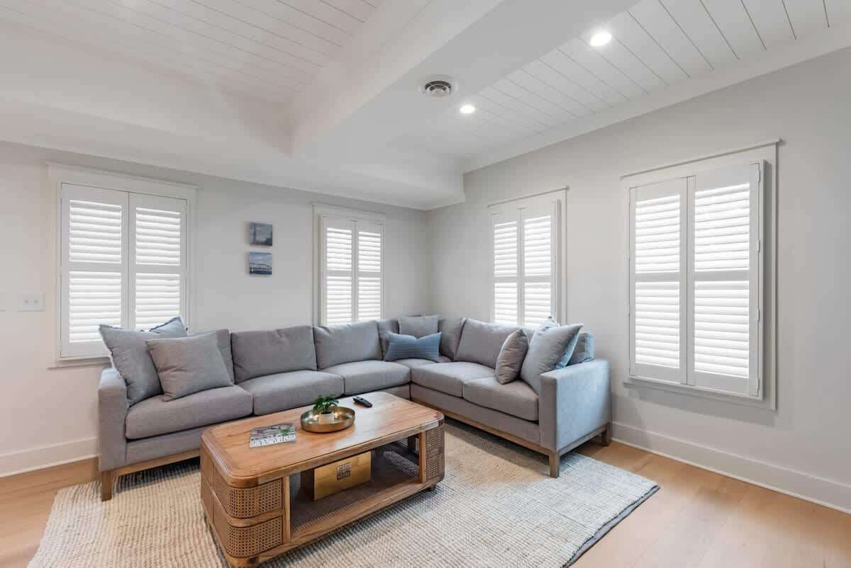A spacious living room with a large L-shaped gray sofa, a wooden coffee table, and white shutters on the windows.