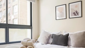 Una cama bien hecha con una colcha a rayas y toallas enrolladas, un ventanal con vistas a edificios, y dos cuadros en la pared.