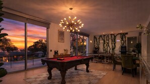 Ein modernes Wohnzimmer mit Billardtisch, großen Fenstern und einer dekorativen Deckenleuchte.